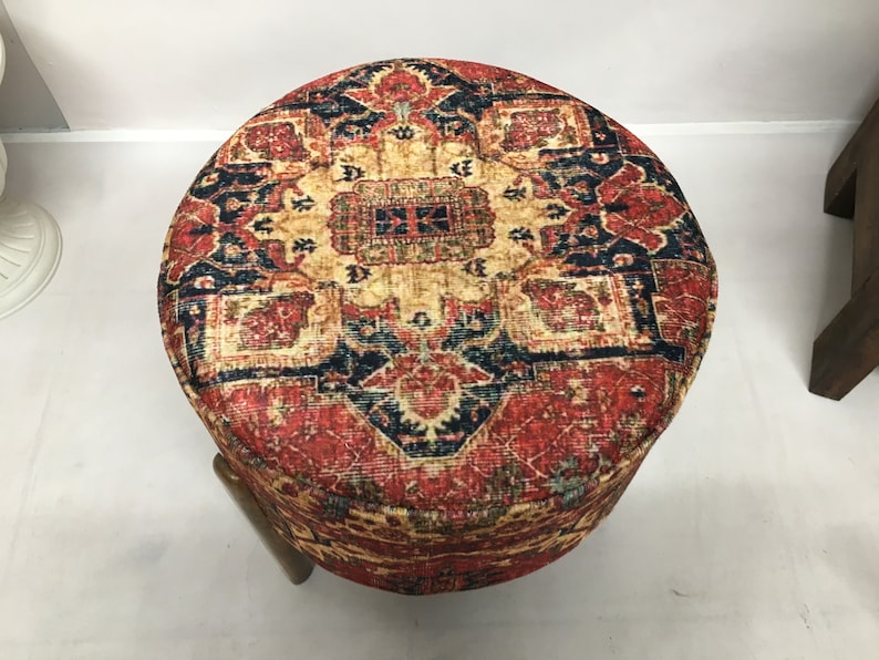 May include: A round ottoman with a colourful, patterned fabric top. The fabric features a red, blue and yellow design with intricate details. The ottoman has a wooden base.