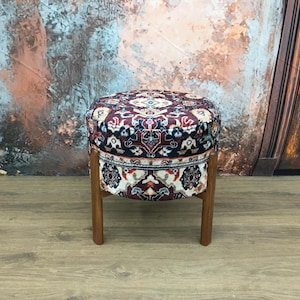 Puede incluir: Un puf redondo con estampado de color rojo oscuro, azul y blanco. El puf tiene cuatro patas de madera.