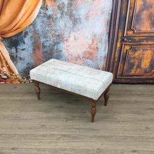 Può includere: Una panca imbottita bianca con un piano in tessuto a fantasia e quattro gambe in legno. La panca è appoggiata su un pavimento in legno davanti a un muro con una finitura di vernice usurata.