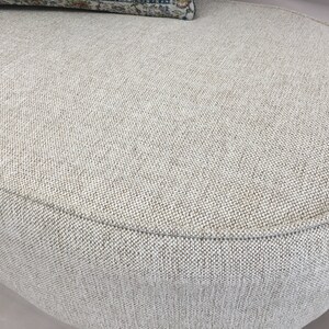 May include: An oval ottoman upholstered in a textured, light beige woven fabric with a visible cross-hatch pattern. The piece features a rounded edge with piping detail along the top perimeter and a small portion of a patterned cushion visible.