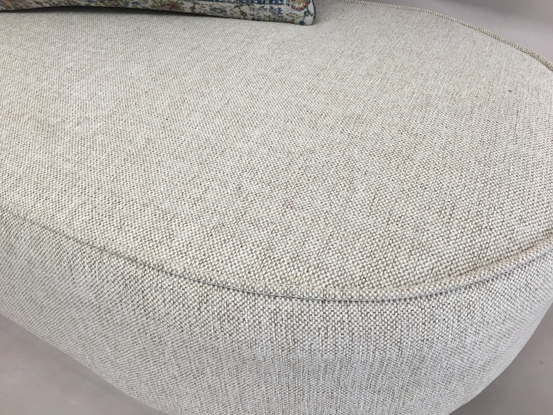 May include: An oval ottoman upholstered in a textured, light beige woven fabric with a visible cross-hatch pattern. The piece features a rounded edge with piping detail along the top perimeter and a small portion of a patterned cushion visible.
