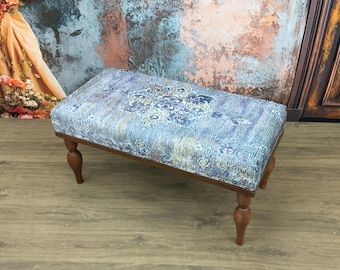 May include: A rectangular bench featuring a cushioned top upholstered in a blue, grey, and cream fabric with a central medallion pattern. The bench is supported by four turned wooden legs with a warm brown finish and a smooth, polished texture.