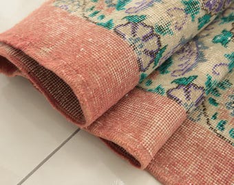 May include: A folded textile rug featuring a wide, solid coral-pink border and a central cream-colored field decorated with a repeating floral pattern in shades of teal, purple, and soft pink, showcasing a visible woven texture and knotted fringe.