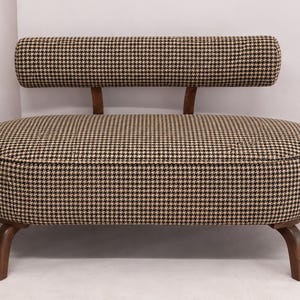 May include: A small bench with a rounded, oval-shaped seat upholstered in a brown and black houndstooth fabric. It features a matching cylindrical backrest supported by two wooden brackets and stands on four curved wooden legs.