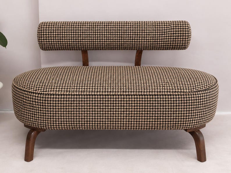 May include: A small bench with a rounded, oval-shaped seat upholstered in a brown and black houndstooth fabric. It features a matching cylindrical backrest supported by two wooden brackets and stands on four curved wooden legs.