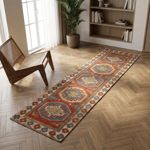 May include: A long rectangular runner rug featuring a repeating geometric pattern in shades of rust red, muted blue, and cream. The design consists of central octagonal medallions surrounded by smaller floral motifs and a decorative border.