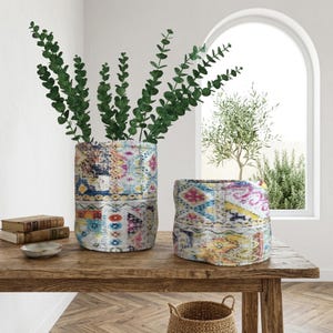 May include: Two fabric vases with vibrant geometric patterns. One is tall and cylindrical, holding green leafy branches. The other is shorter and wider. They sit on a wooden table. A small bowl and books are also on the table.