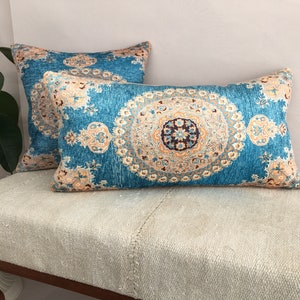 May include: Two decorative throw pillows with a blue background and intricate patterns in shades of beige, orange, and brown. The pillows are on a light beige couch.