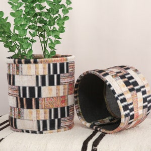 Könnte beinhalten: Zwei zylindrische Stoffaufbewahrungsbehälter mit einem geometrischen Muster in Schwarz, Weiß und Beige. Der Behälter links enthält eine Topfpflanze mit grünen Blättern.