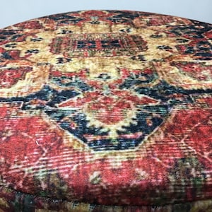 May include: Close-up of a round, red and gold patterned ottoman with a velvet-like texture. The pattern features intricate details and a central design with a dark blue and red colour scheme.