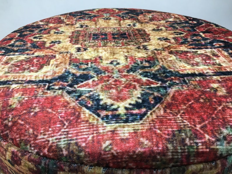 May include: Close-up of a round, red and gold patterned ottoman with a velvet-like texture. The pattern features intricate details and a central design with a dark blue and red colour scheme.