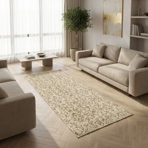 May include: A beige rectangular rug with a detailed floral pattern is laid on a wooden floor in a living room setting. The rug is positioned between two beige sofas. A coffee table and a potted tree are also visible.
