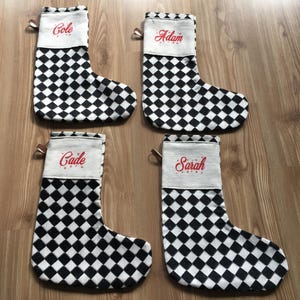 Puede incluir: Cuatro calcetines de Navidad blancos con patrones de cuadros negros y nombres bordados en rojo: Cole, Adam, Cade y Sarah.