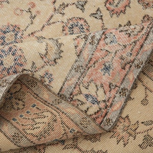 May include: Close-up of a beige rug with a floral pattern. The rug has a light pink and blue border. The rug is folded in half, showing the underside.