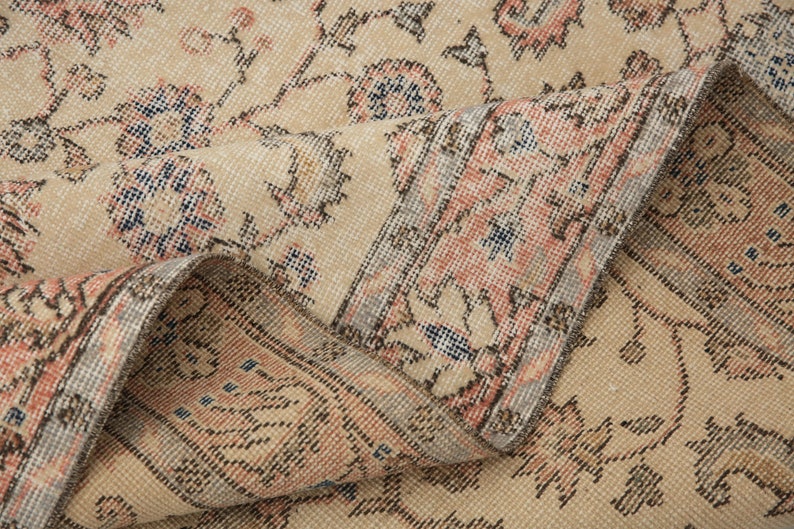 May include: Close-up of a beige rug with a floral pattern. The rug has a light pink and blue border. The rug is folded in half, showing the underside.