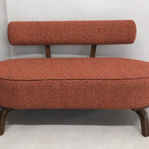 May include: A small, oval-shaped loveseat upholstered in a textured, burnt orange fabric. The piece features a low, floating backrest supported by two wooden brackets and is elevated on four curved, dark brown wooden legs.