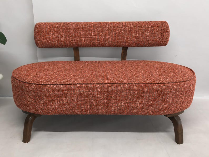 May include: A small, oval-shaped loveseat upholstered in a textured, burnt orange fabric. The piece features a low, floating backrest supported by two wooden brackets and is elevated on four curved, dark brown wooden legs.