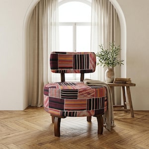 Puede incluir: Una silla decorativa de patchwork con respaldo bajo y patas de madera oscura. El asiento y el respaldo presentan una colorida mezcla de patrones geométricos en tonos rojos, naranjas, rosas y negros. Una manta de color neutro está sobre el asiento.