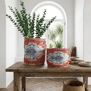 May include: Two cylindrical planters with a red, blue, and white patterned design sit on a wooden table. The larger planter holds green leafy branches. The smaller planter is empty. The table is in front of a window.