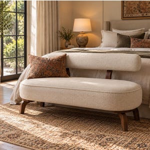 May include: An off-white upholstered bench with a rounded seat and a matching cylindrical backrest supported by dark wooden legs. A rectangular throw pillow with a dark orange and brown patterned design rests on the seat.