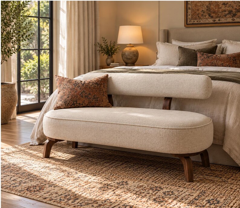 May include: An off-white upholstered bench with a rounded seat and a matching cylindrical backrest supported by dark wooden legs. A rectangular throw pillow with a dark orange and brown patterned design rests on the seat.