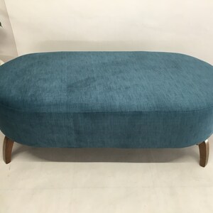 May include: A teal blue oval ottoman with wooden legs. The ottoman is upholstered in a soft, textured fabric.