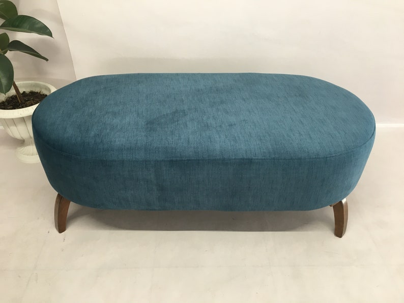 May include: A teal blue oval ottoman with wooden legs. The ottoman is upholstered in a soft, textured fabric.