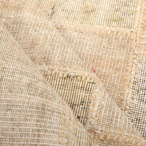 Puede incluir: Una alfombra de patchwork en tonos beige y marrón claro que presenta una cuadrícula de secciones rectangulares de tela unidas por costuras visibles. El tejido muestra una textura trenzada con tonos crema y beige, con una esquina doblada.