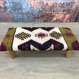 May include: A small, rectangular ottoman with wooden legs. The top is covered in a colourful, patterned fabric with geometric designs in shades of red, green, black, and white.