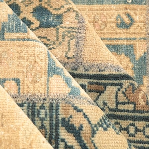 May include: Close-up of a folded rug with a beige background and blue and brown geometric patterns.