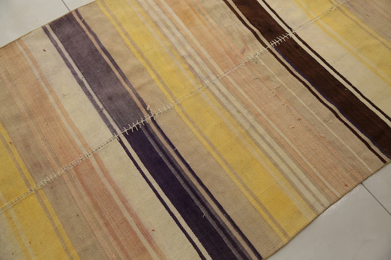 May include: A rectangular rug with a patchwork design. The rug features alternating stripes of yellow, beige, brown, and purple. The stripes run diagonally across the rug. The rug is made of a woven material.