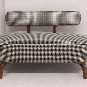 May include: A small loveseat featuring a black and white houndstooth fabric upholstery. The piece has a rounded, oval-shaped seat, a low-profile cylindrical backrest supported by two wooden brackets, and four curved wooden legs.