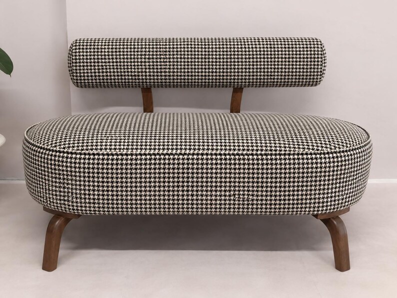 May include: A small loveseat featuring a black and white houndstooth fabric upholstery. The piece has a rounded, oval-shaped seat, a low-profile cylindrical backrest supported by two wooden brackets, and four curved wooden legs.