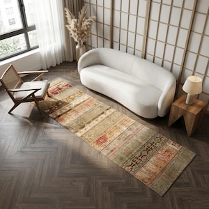 May include: A rectangular, distressed-look rug with a patchwork design in shades of beige, red, and brown. The rug is placed on a dark wood floor in a living room setting, alongside a white sofa and a wooden chair.