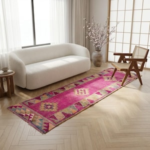 May include: A long, rectangular, magenta-colored rug with geometric patterns in shades of yellow, blue, and green. The rug is placed on a light-colored wooden floor, with a white sofa and wooden chair in the background. The rug adds a pop of color to the room.