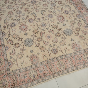 May include: A vintage rug with a cream background and a floral pattern in shades of pink, blue, and gray. The rug has a border with a repeating pattern of flowers and leaves.