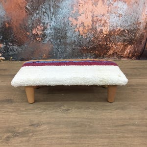 May include: A small, rectangular footstool with a white, textured top and four wooden legs. The top features a stripe of red, blue, and brown fabric. The stool is designed for comfort and adds a touch of rustic charm to any space.
