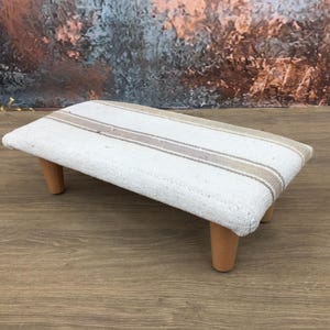 May include: A small, rectangular footstool with a white fabric top featuring tan stripes. The stool has four short, light-colored wooden legs. The background is a blurred mix of colors.