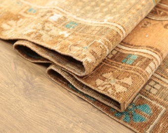 May include: A woven area rug featuring a textured pile with a geometric pattern in shades of tan, brown, and beige, accented by small, repeating floral-like motifs in a vibrant turquoise blue color throughout the border and central field.