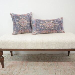 May include: A white upholstered bench with a wooden frame and turned legs. The bench has two patterned pillows on top, one square and one rectangular.