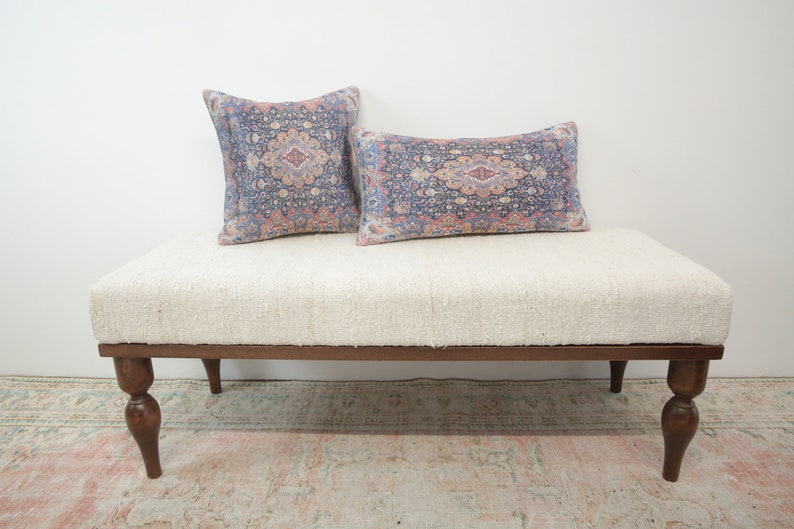 May include: A white upholstered bench with a wooden frame and turned legs. The bench has two patterned pillows on top, one square and one rectangular.
