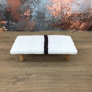 May include: A small, rectangular white footstool with a black stripe down the center. The stool is supported by four short, light-colored wooden legs. The footstool is placed on a wooden surface.
