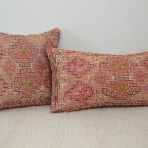 May include: Two decorative throw pillows featuring a geometric pattern in shades of pink, red, and yellow. The pillows are made from woven fabric and have a fringed edge.