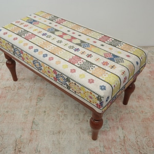 Pode incluir: Banqueta retangular com tampo de tecido estampado com cores como amarelo, vermelho, azul e branco. A banqueta tem quatro pernas torneadas em castanho escuro. O design sugere um estilo artesanal ou vintage, adequado para decoração de interiores. Mede cerca de 90 cm de comprimento.