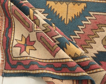 May include: A woven rug featuring a geometric pattern in shades of blue, dusty rose, mustard yellow, and cream. The rug has a folded corner revealing a fringed edge and a repeating design of stars, zigzags, and angular shapes across the surface.