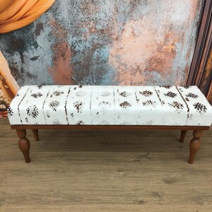 May include: A white bench with a brown and white patterned cushion and wooden legs. The bench is sitting on a wooden floor.