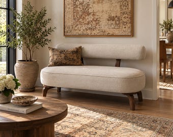 May include: A cream-coloured upholstered bench with a rounded backrest and seat, supported by dark wooden legs. A rectangular brown patterned throw pillow rests on the seat. The bench features a clean, simple design with visible fabric texture.