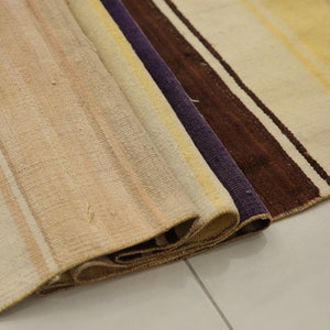 May include: A rolled-up, striped rug with a neutral color palette. The rug features vertical stripes in shades of beige, cream, yellow, brown, and purple. The rug's texture is visible, and the edges are slightly frayed.