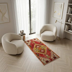 Può includere: Un interno con due poltrone arrotondate color crema e un vivace tappeto rettangolare. Il tappeto ha uno sfondo rosso con un disegno geometrico centrale giallo e verde e motivi floreali. La stanza ha una palette di colori neutri con una finestra e una libreria.
