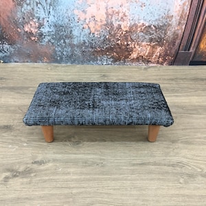 May include: A small, rectangular footstool with a dark blue and white patterned top. The stool has four short, light brown wooden legs. The background features a wooden floor and a wall with a faded, abstract design.